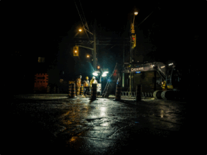 Construction workers and a Deere excavator work at a road site at night, illuminated by streetlights and surrounded by traffic cones.