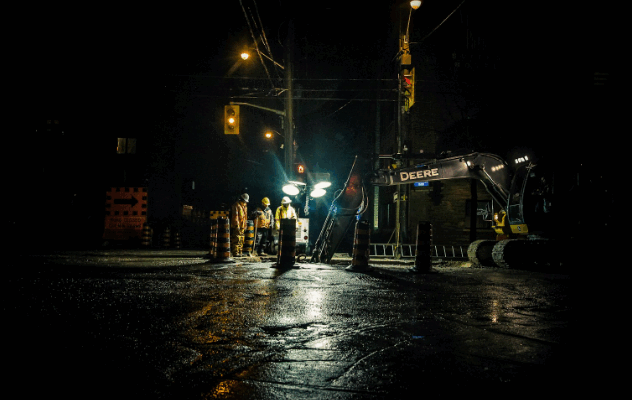 Construction workers and a Deere excavator work at a road site at night, illuminated by streetlights and surrounded by traffic cones.
