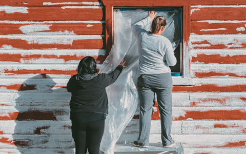 Two people are covering a window with clear plastic sheeting on the exterior of a red and white wooden building.
