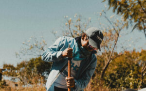 Person wearing a cap and denim jacket works outdoors with a wooden tool, surrounded by trees and a clear sky.