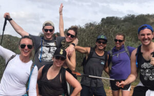 A group of seven people wearing casual outdoor clothing pose and smile for a photo while hiking on a trail, surrounded by shrubs and hills.