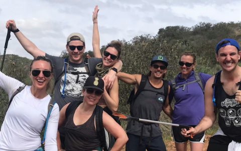 A group of seven people wearing casual outdoor clothing pose and smile for a photo while hiking on a trail, surrounded by shrubs and hills.