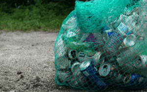 A green net bag filled with empty aluminum cans sits on a gravel surface near some grass.