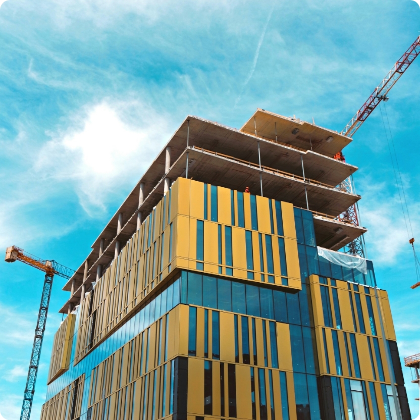 A modern building under construction with yellow and blue patterned glass panels, cranes, and workers visible against a blue sky.