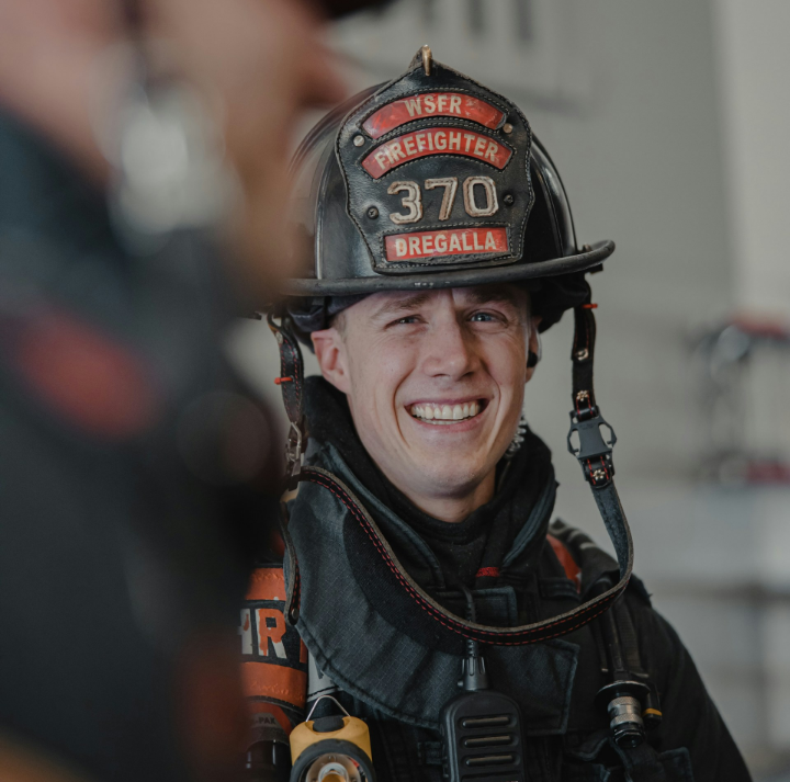 A firefighter in uniform and helmet smiles indoors, with firefighting equipment visible in the background.