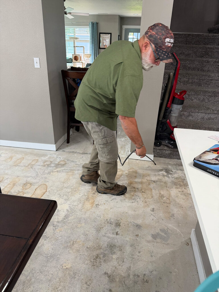 A man in a green shirt and cap removes old flooring using a tool in a home, with furniture and a vacuum visible in the background.