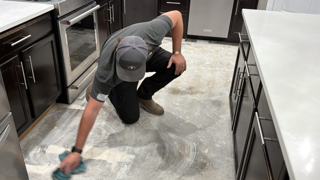 A person wearing work clothes is kneeling on the floor of a kitchen, cleaning the bare concrete surface with a cloth.
