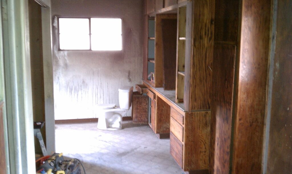 A bathroom with wooden cabinets, a toilet, dirty walls, and a window; the room appears worn and neglected.