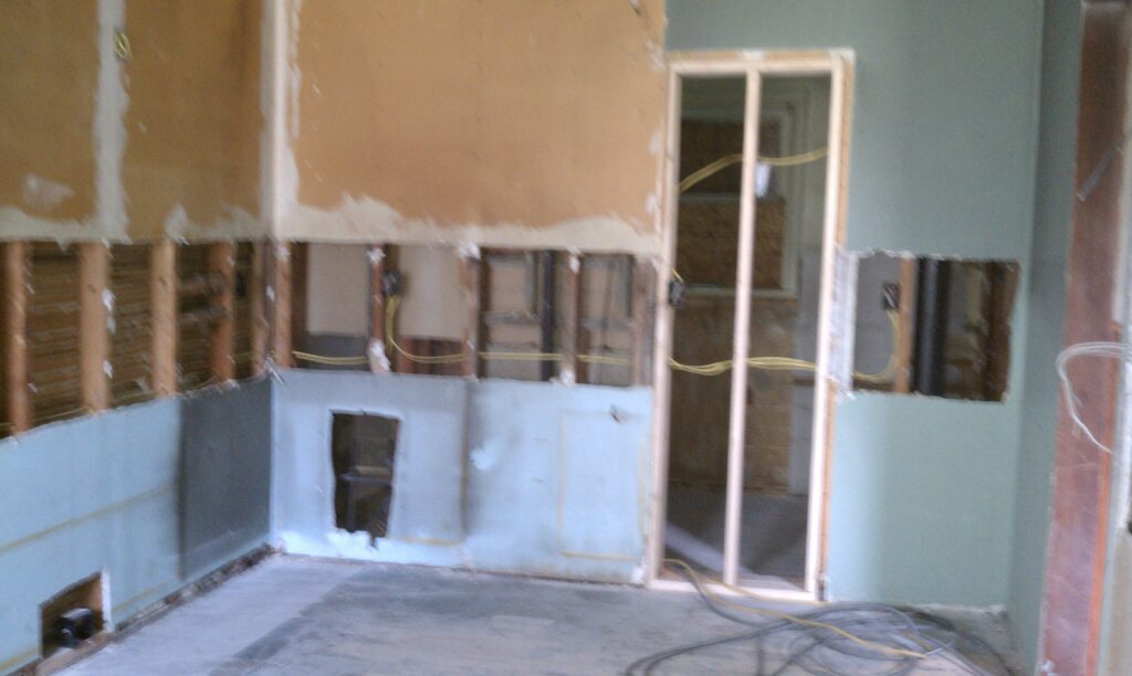 Partially demolished room with exposed wall studs, electrical wiring, missing drywall, and construction materials on the floor.