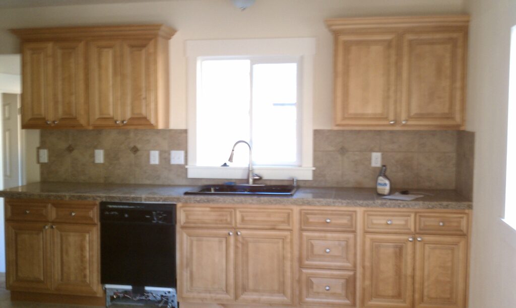 Kitchen with light wood cabinets, granite countertop, stainless steel sink, dishwasher, and tiled backsplash. A bottle is placed near the right side of the sink.