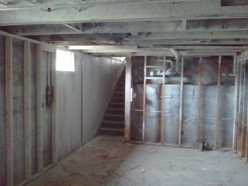 Unfinished basement with exposed wooden studs, insulation, concrete floor, and a staircase leading up. A small window lets in light.