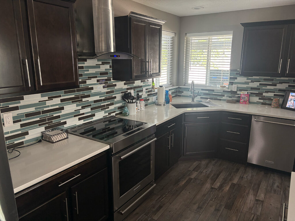 Modern kitchen with dark cabinets, stainless steel appliances, white countertops, and glass tile backsplash. Sunlight streams in through two windows above the sink.