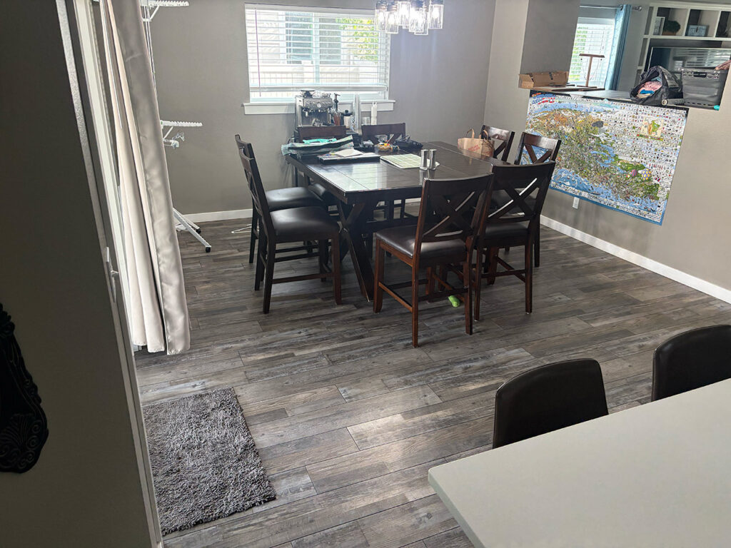 Dining room with a square table and six chairs, puzzle pieces on the table, a large puzzle on a side table, and bright daylight coming through two windows.