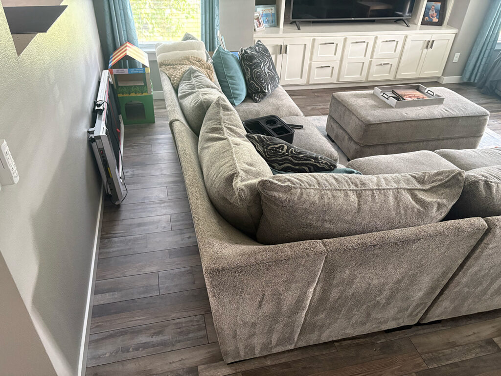 A gray sectional sofa with blue and patterned pillows is in a living room with wood floors, a TV, a large ottoman, and children’s toys near the window.