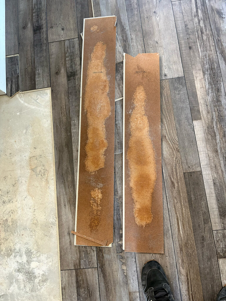 Two damaged planks of laminate flooring with visible water stains are placed on a floor with matching undamaged planks; part of a shoe is visible at the bottom.