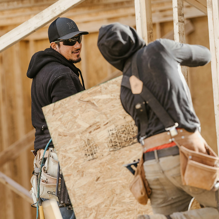 Two construction workers wearing tool belts and hoodies handle a sheet of plywood at a building site framed with wooden beams.