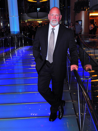 A man in a suit stands on a blue-lit glass walkway indoors, resting one hand on a railing, with people and lights visible in the background.