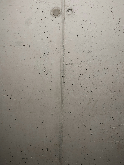 A plain concrete wall with a vertical seam down the center, small holes, and scattered dark spots throughout the surface.