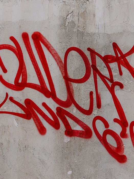 Red spray-painted graffiti on a rough, weathered concrete wall. The writing is stylized and illegible.