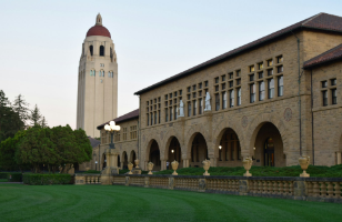 A large stone building with arched windows and a red-roofed tower in the background, surrounded by manicured lawns and trees.